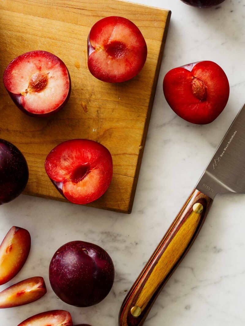 A wooden cutting board with halved fresh black plums and a knife. 