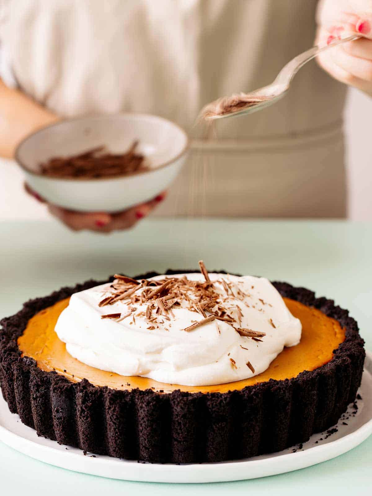 A whole pumpkin pie with chocolate crust and whipped cream with shaved chocolate being sprinkled on.