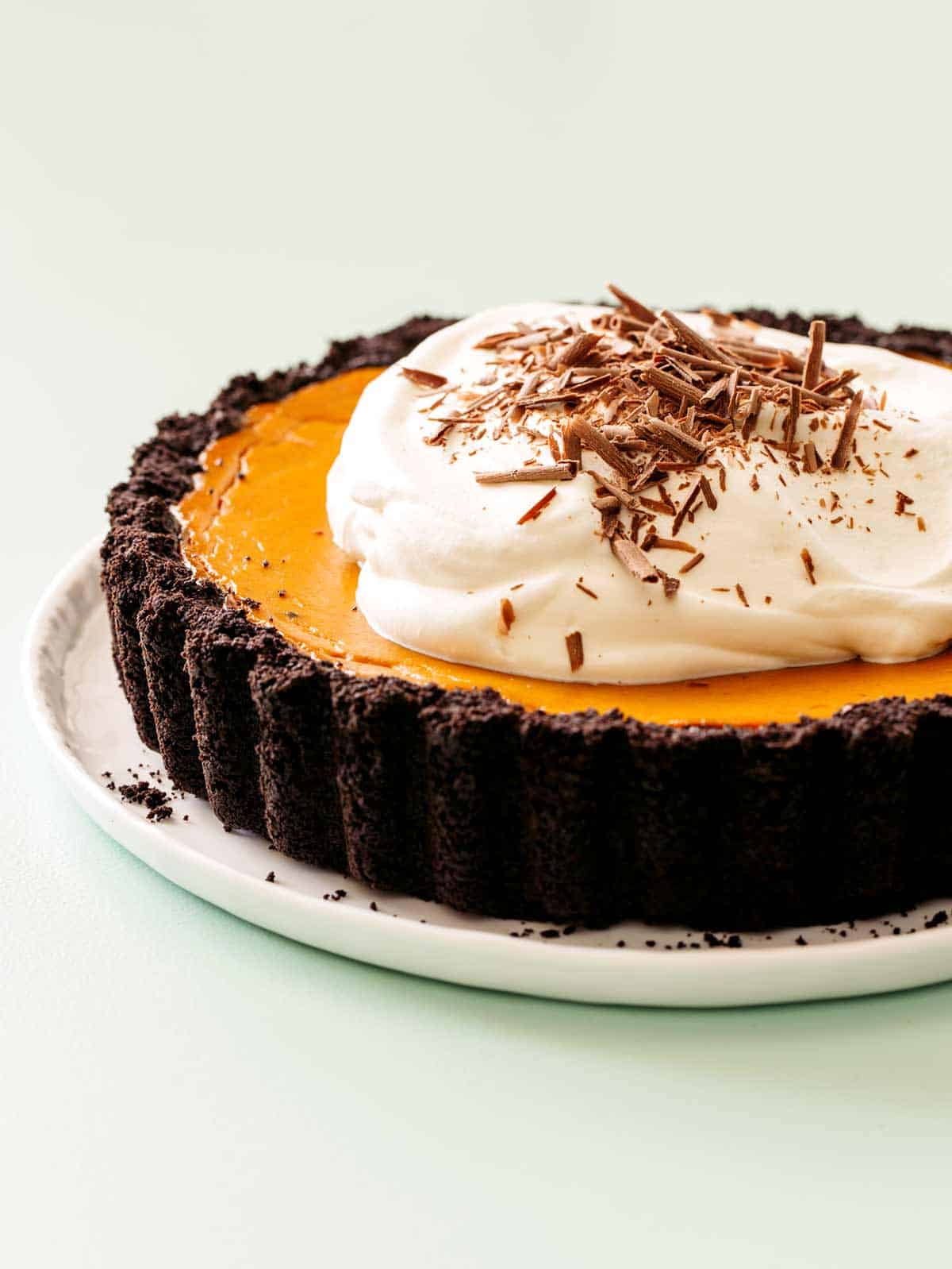 A close up of a whole pumpkin pie with chocolate crust, whipped cream, and chocolate shavings.