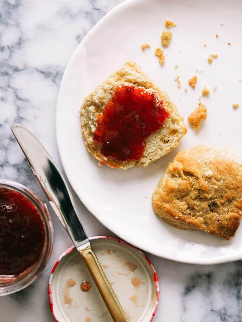 A halved brown butter biscuit covered in jam, with a jar of jam and knife.