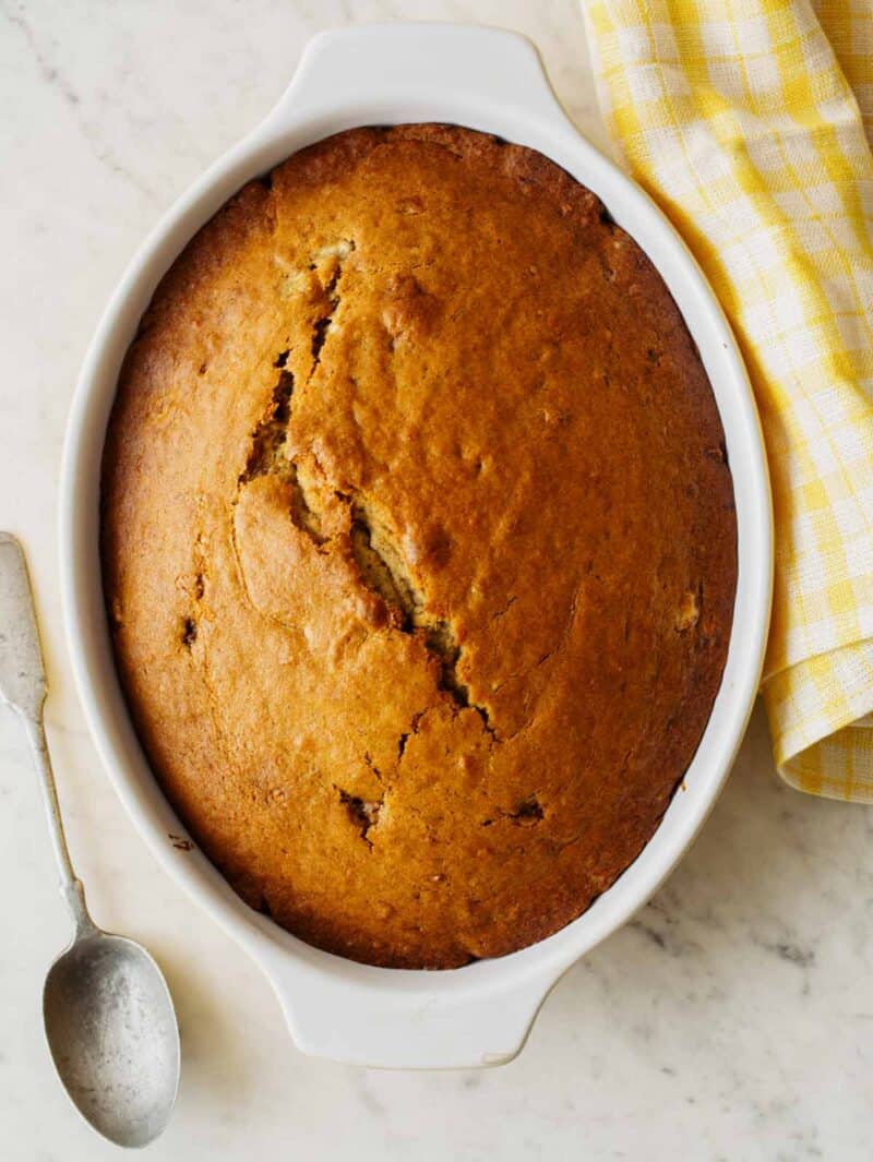 A whole chai banana cake in an oval baking dish with a spoon and linen.