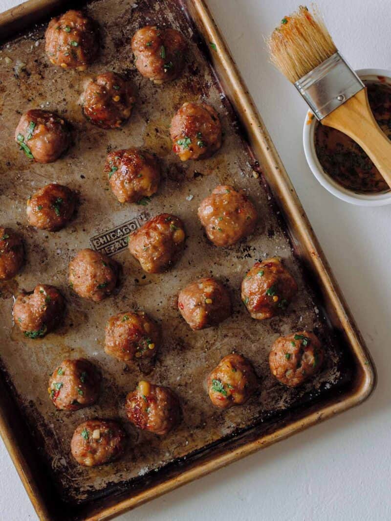 A sheet pan of finished ginger garlic cocktail meatballs with a bowl of glaze and brush.