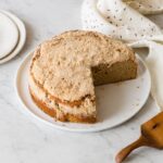 A sour cream and cinnamon crumb cake with a piece taken out and plates and linens.