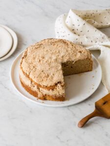 A sour cream and cinnamon crumb cake with a piece taken out and plates and linens.
