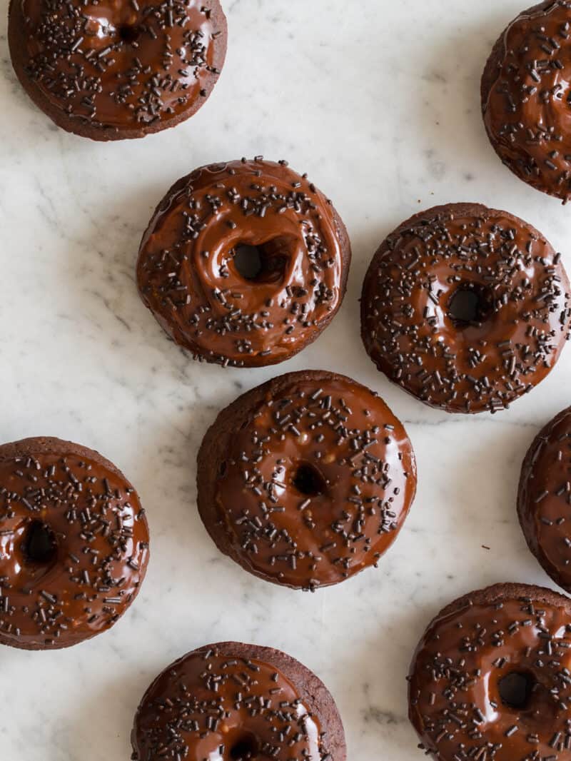 Baked chocolate doughnuts with chocolate glaze and chocolate sprinkles on a marble surface.