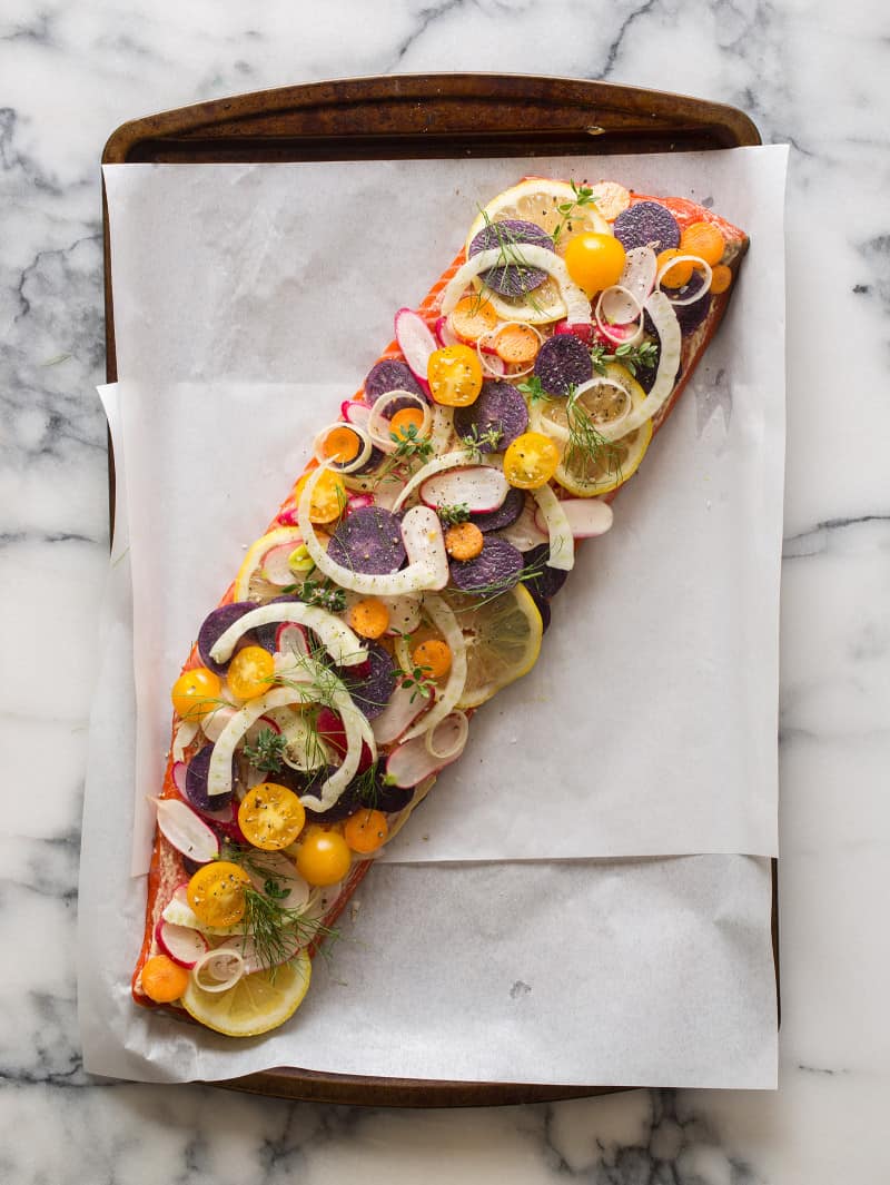 A whole side of salmon stacked with veggies and herbs on parchment paper.