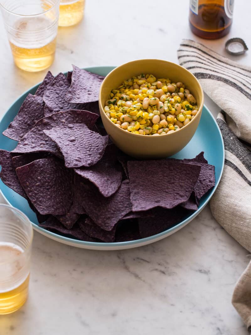 A bowl of white bean and corn salsa on a plate of blue corn tortilla chips.