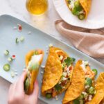 A person taking a taco off a plate of spicy curry shrimp tacos with a drink.