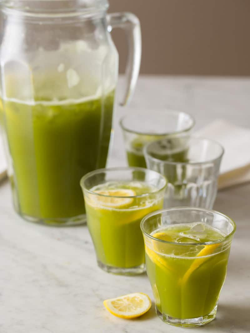 A glass pitcher of matcha lemonade with several full glasses and lemon slices.