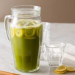 A glass pitcher of matcha lemonade with extra glasses and lemon slices.