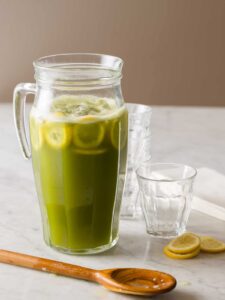 A glass pitcher of matcha lemonade with extra glasses and lemon slices.