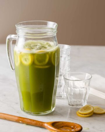 A glass pitcher of matcha lemonade with extra glasses and lemon slices.