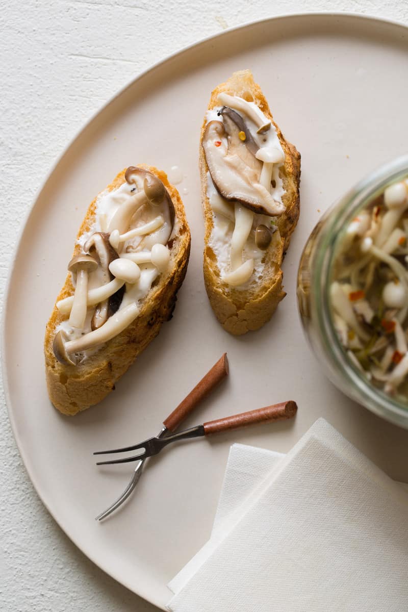 Two crostinis with pickled mushrooms on top, and a jar of pickled mushrooms next to them. 