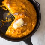 Pumpkin cornbread in a cast iron skillet with a piece cut out and butter on top.