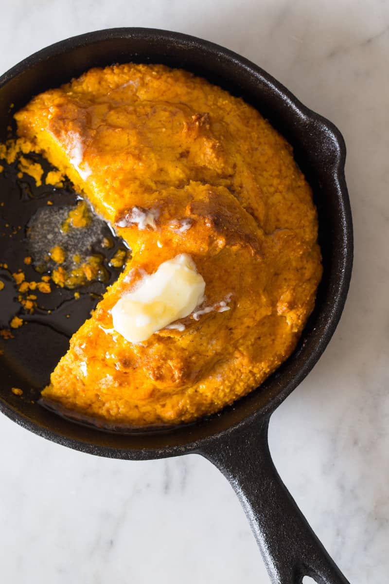 A close up of pumpkin cornbread with butter in a skillet and a piece cut out.