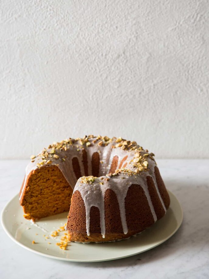 A pumpkin bundt cake topped with pistachio glaze and pistachios with a piece cut out.