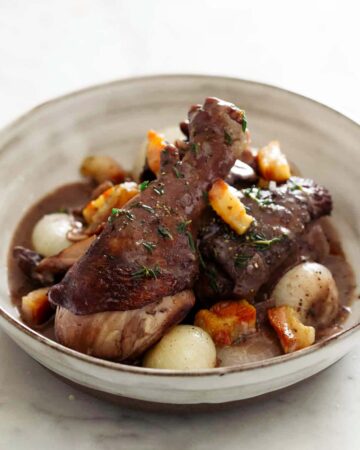 A close up of a bowl of coq au vin with bread, a fork, and a knife.