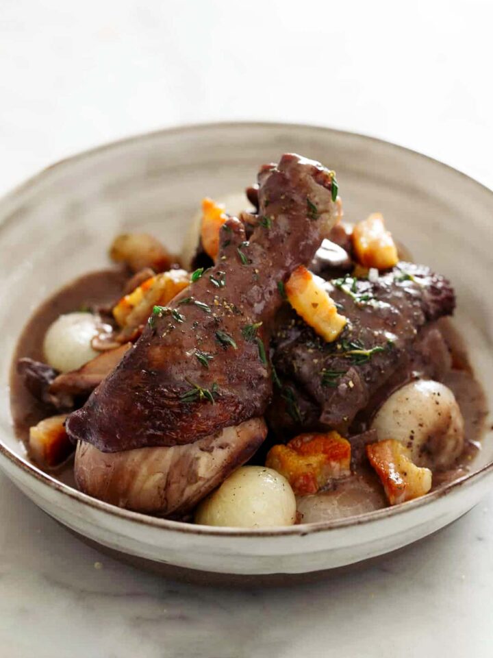 A close up of a bowl of coq au vin with bread, a fork, and a knife.