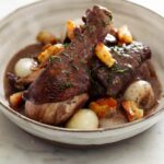 A close up of a bowl of coq au vin with bread, a fork, and a knife.