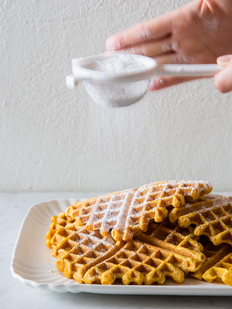 Stacked pumpkin spiced waffles with powdered sugar being sifted on top.