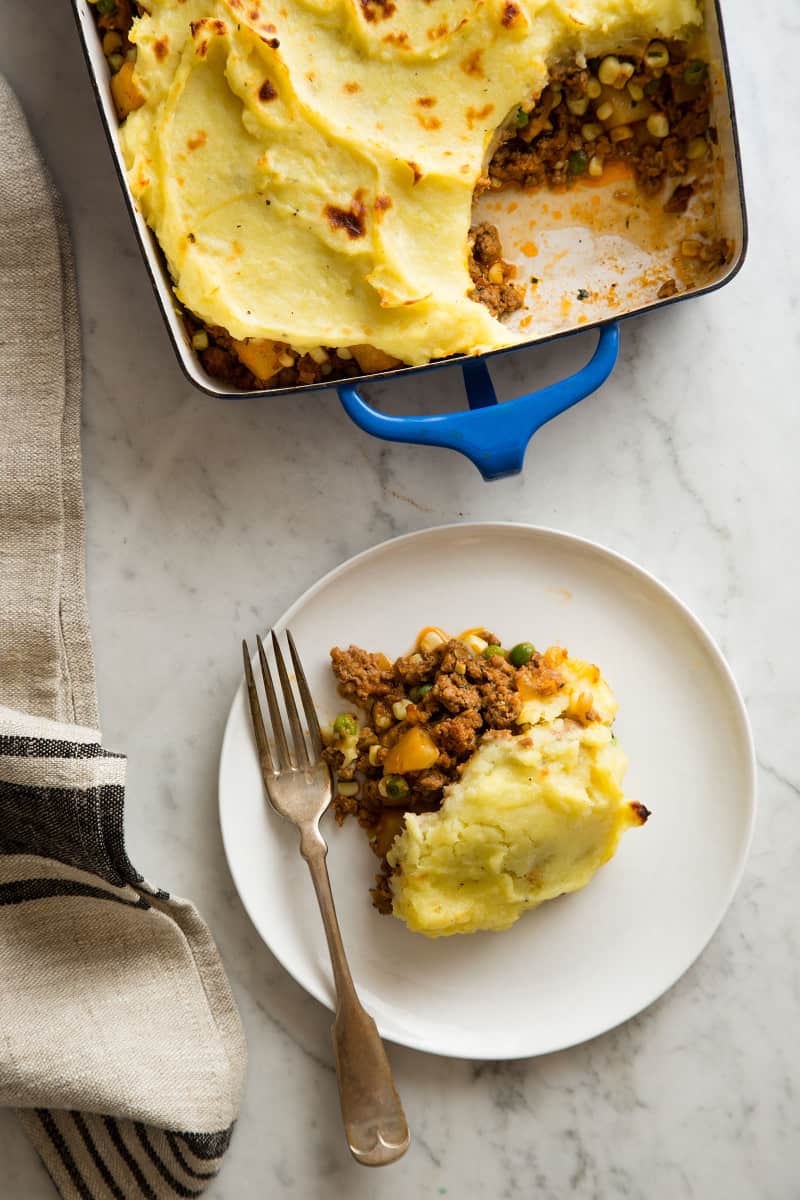 A casserole dish of shephers pie with a scoop out on a plate. 