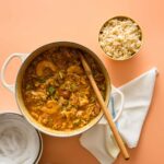 A pot of chicken and andouille gumbo with a bowl of rice and a wooden spoon.