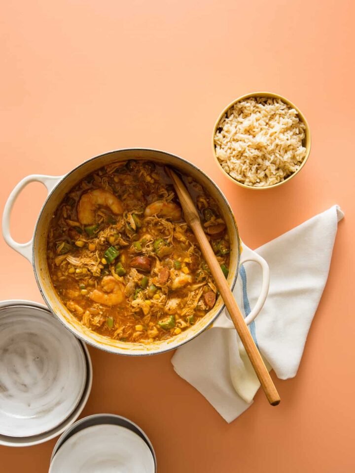 A pot of chicken and andouille gumbo with a bowl of rice and a wooden spoon.