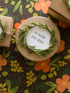 A close up of a rosemary wreath gift toppers tied to a round brown gift box.