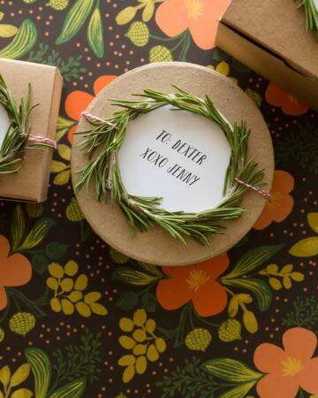 A close up of a rosemary wreath gift toppers tied to a round brown gift box.