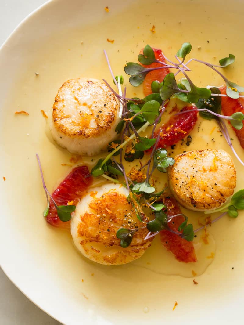 A close up of citrus seared scallops garnished with blood orange segments and micro greens.