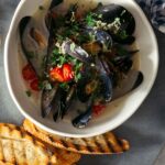 A bowl of white wine steamed mussels on a plate with bread, forks, and drinks.