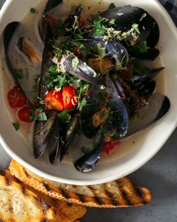 A bowl of white wine steamed mussels on a plate with bread, forks, and drinks.