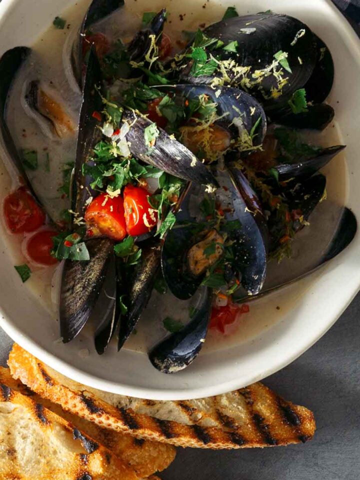 A bowl of white wine steamed mussels on a plate with bread, forks, and drinks.