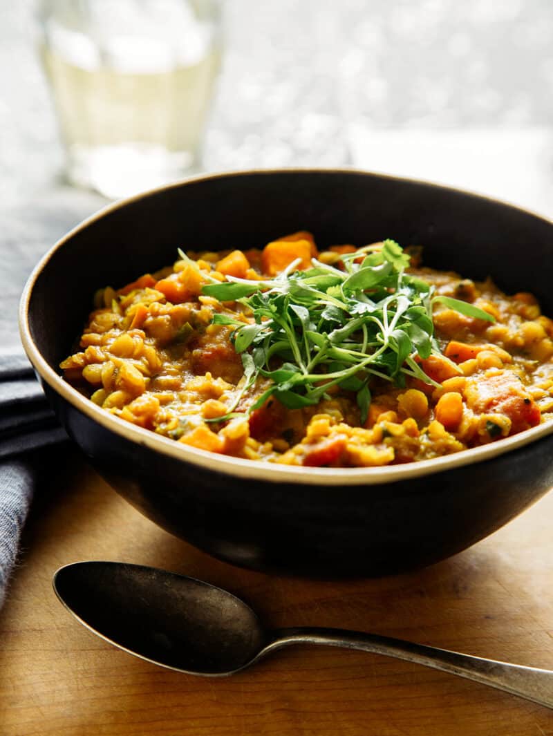 A bowl of mansoor dal with a spoon next to it and a glass of wine in the background.