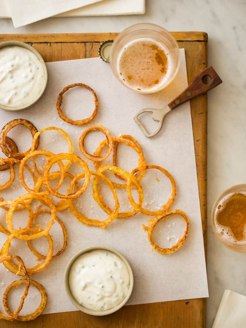 A wooden cutting board with extra crispy beer battered onion rings, beer, and dipping sauce.