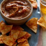 A bowl of roasted tomato salsa on a plate of tortilla chips with a drink.