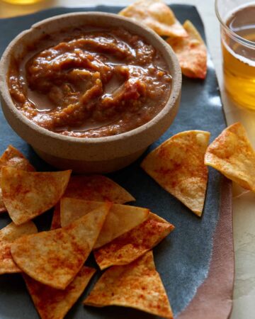 A bowl of roasted tomato salsa on a plate of tortilla chips with a drink.