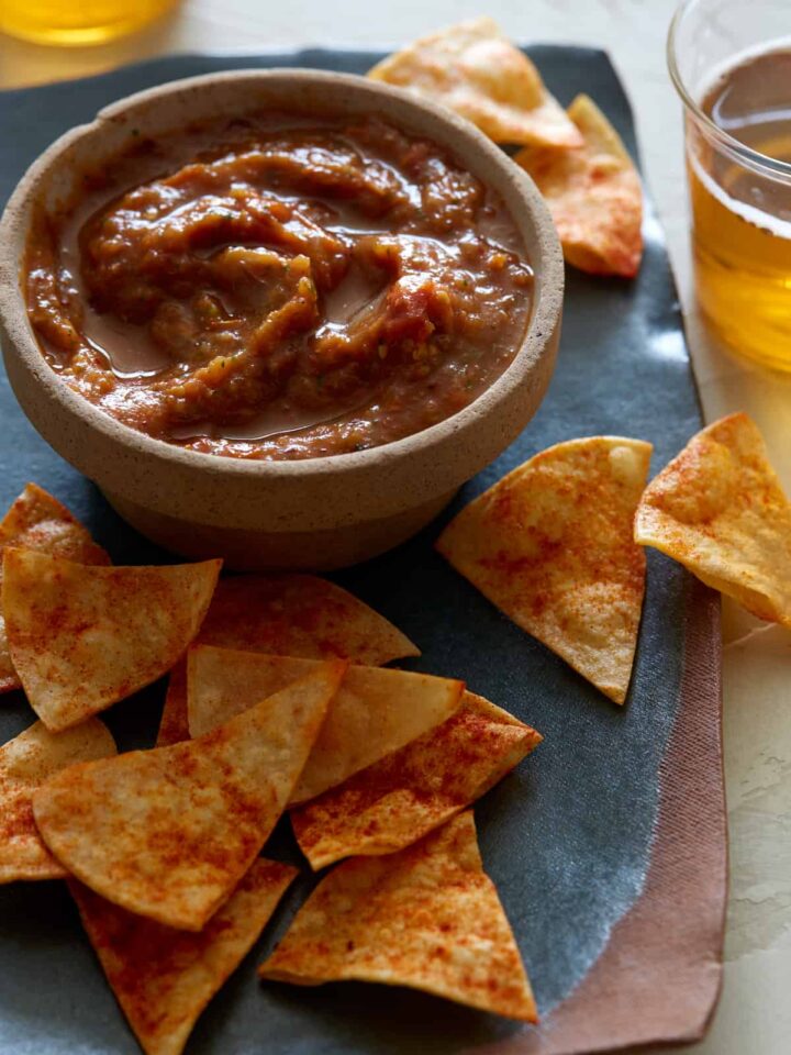 A bowl of roasted tomato salsa on a plate of tortilla chips with a drink.