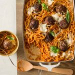 Baked bucatini and meatballs in a baking dish with a bowl served and a wooden spoon.
