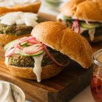 Falafel burgers with veggies and sauce on a wooden cutting board.