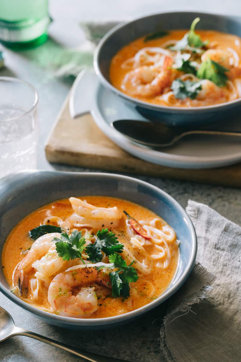 Bowls of spicy red curry and coconut noodle soup with shrimp and a spoon.