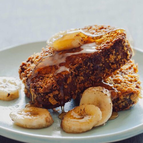 Two pieces of banana bread french toast with caramelized bananas and maple syrup on a plate.