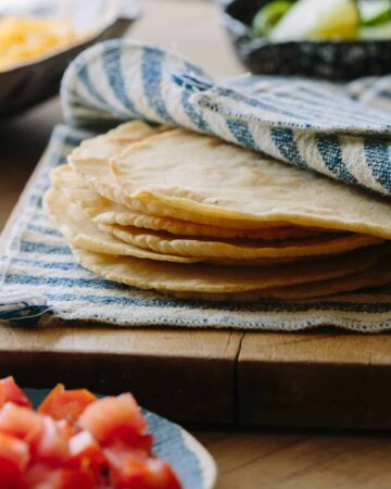 Homemade corn tortillas a hand towel on a wooden cutting board.