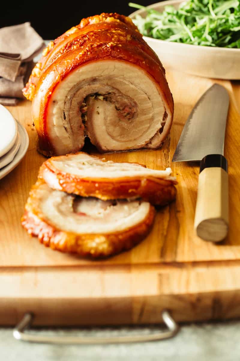 A close up of sliced pork belly porchetta with a knife on a cutting board.