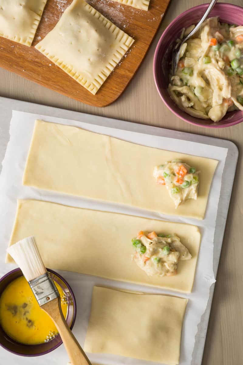 Rolled out, cut dough being filled with chicken pot pie filling, egg and a brush.