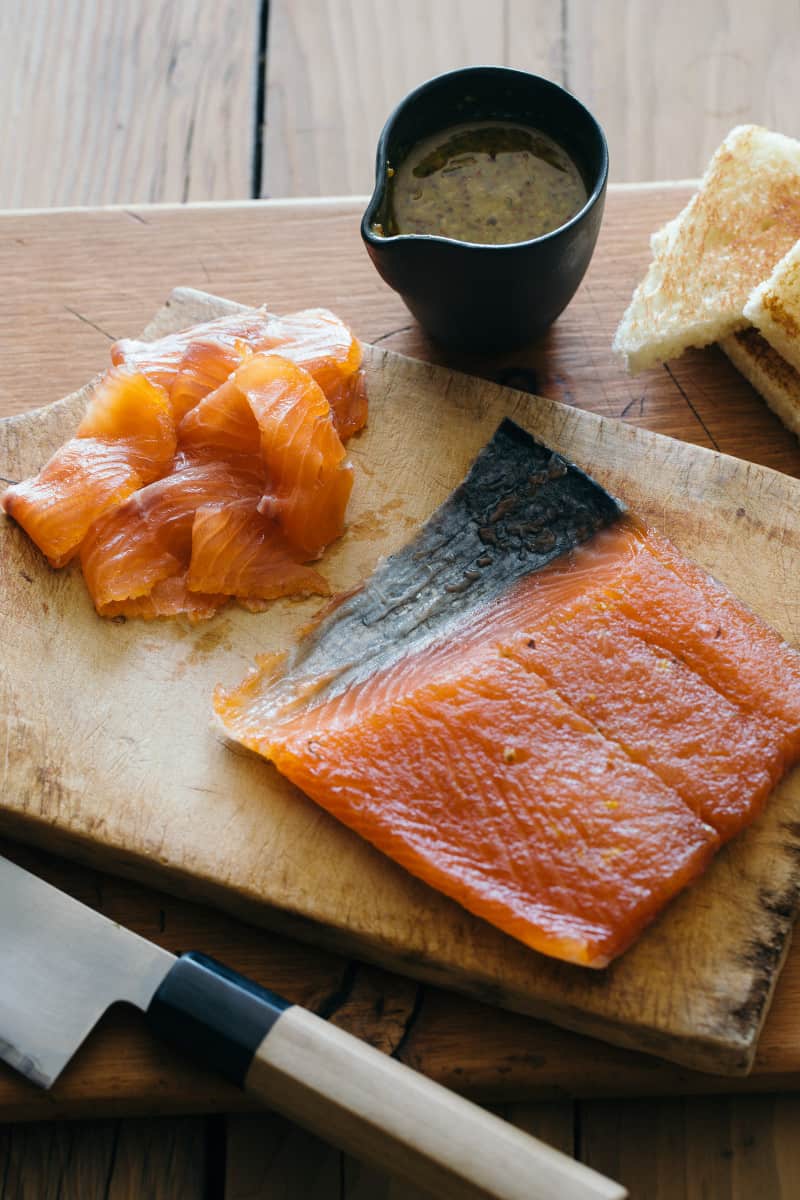 A piece of salmon with slices on a wooden cutting board and sauce on the side.