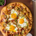A breakfast pizza on a wooden cutting board with a bowl of chives on the side.