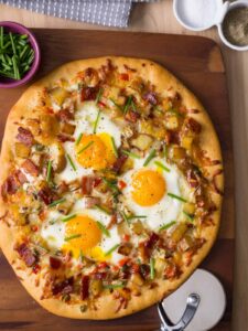 A breakfast pizza on a wooden cutting board with a bowl of chives on the side.