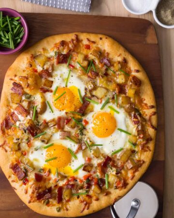 A breakfast pizza on a wooden cutting board with a bowl of chives on the side.