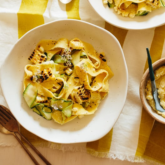 Bowls of sweet corn pesto with pappardelle and zucchini noodles on linens with forks and spoons.